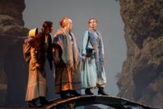Three costumed performers stand hand-in-hand on a curved platform between towering rocky cliffs.