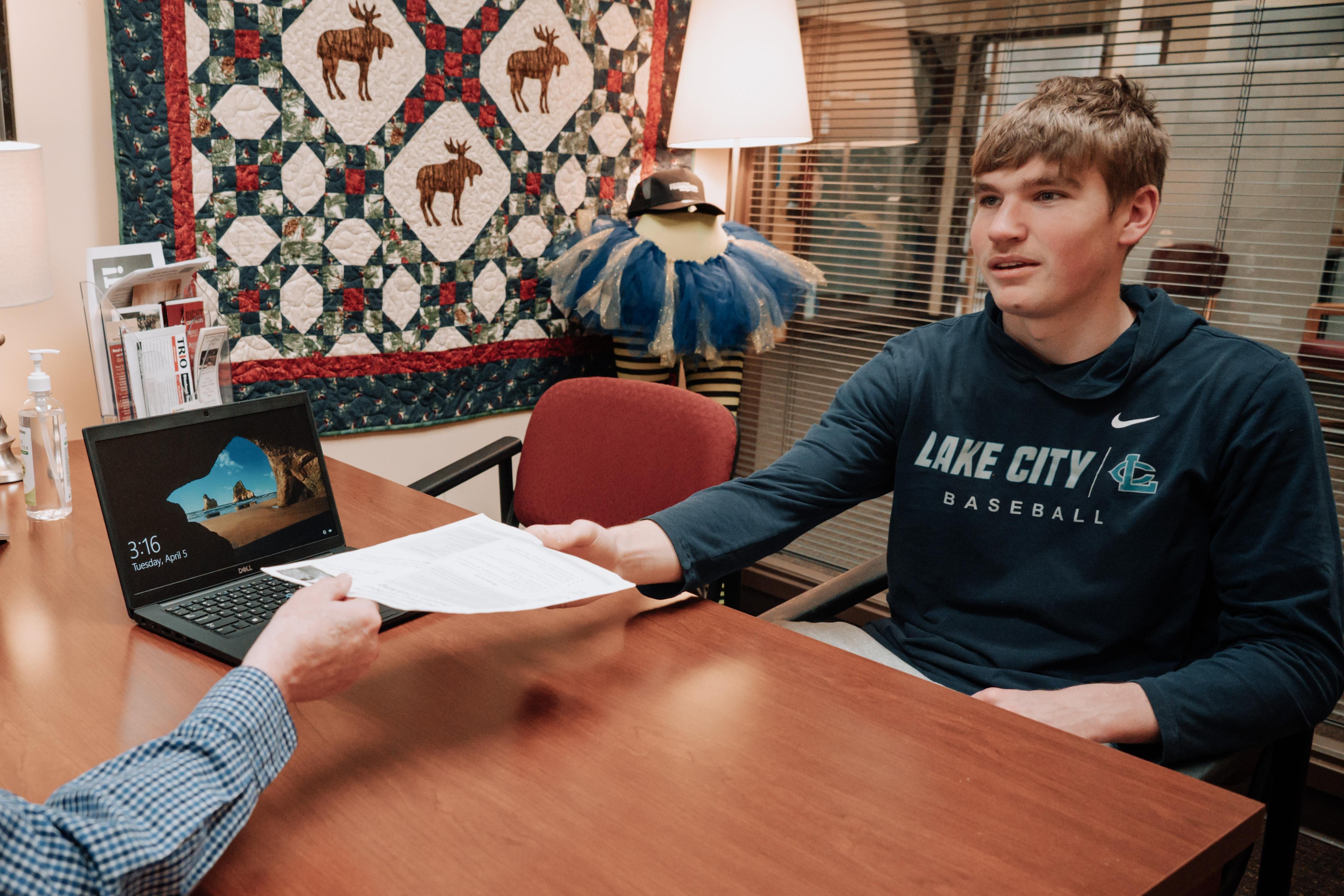 Male student sitting at desk getting paperwork from Instructor