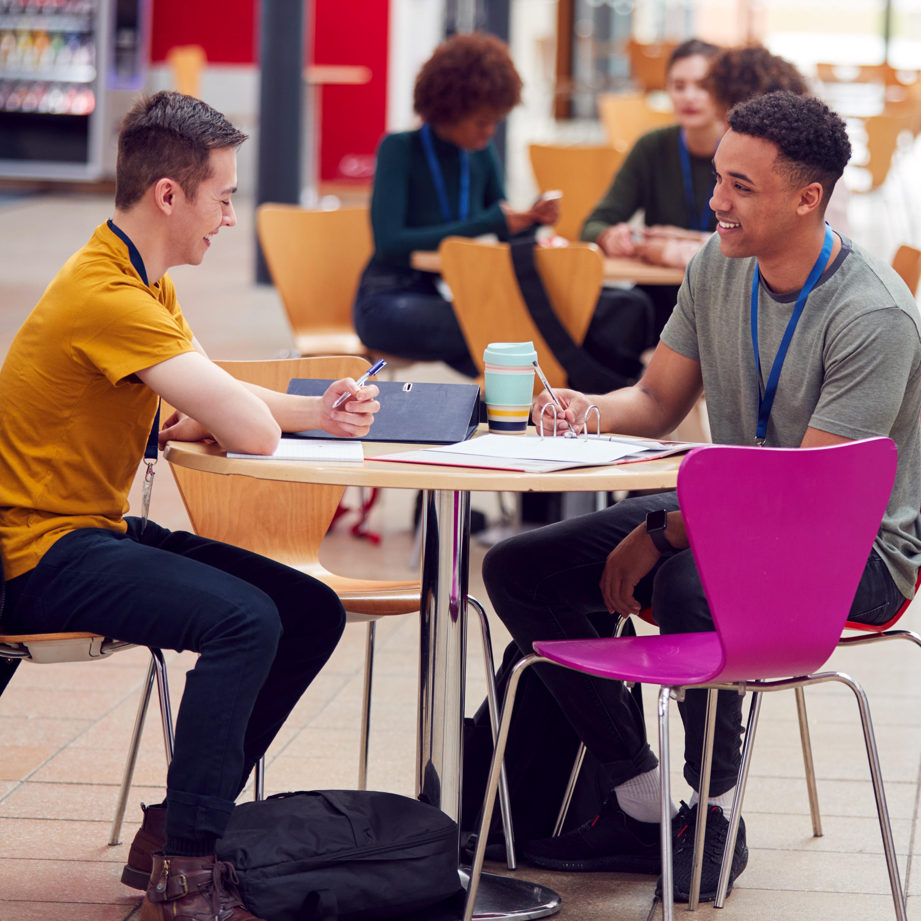 Two young men engaged in conversation at a wooden table in a brightly lit university cafeteria, surrounded by other students