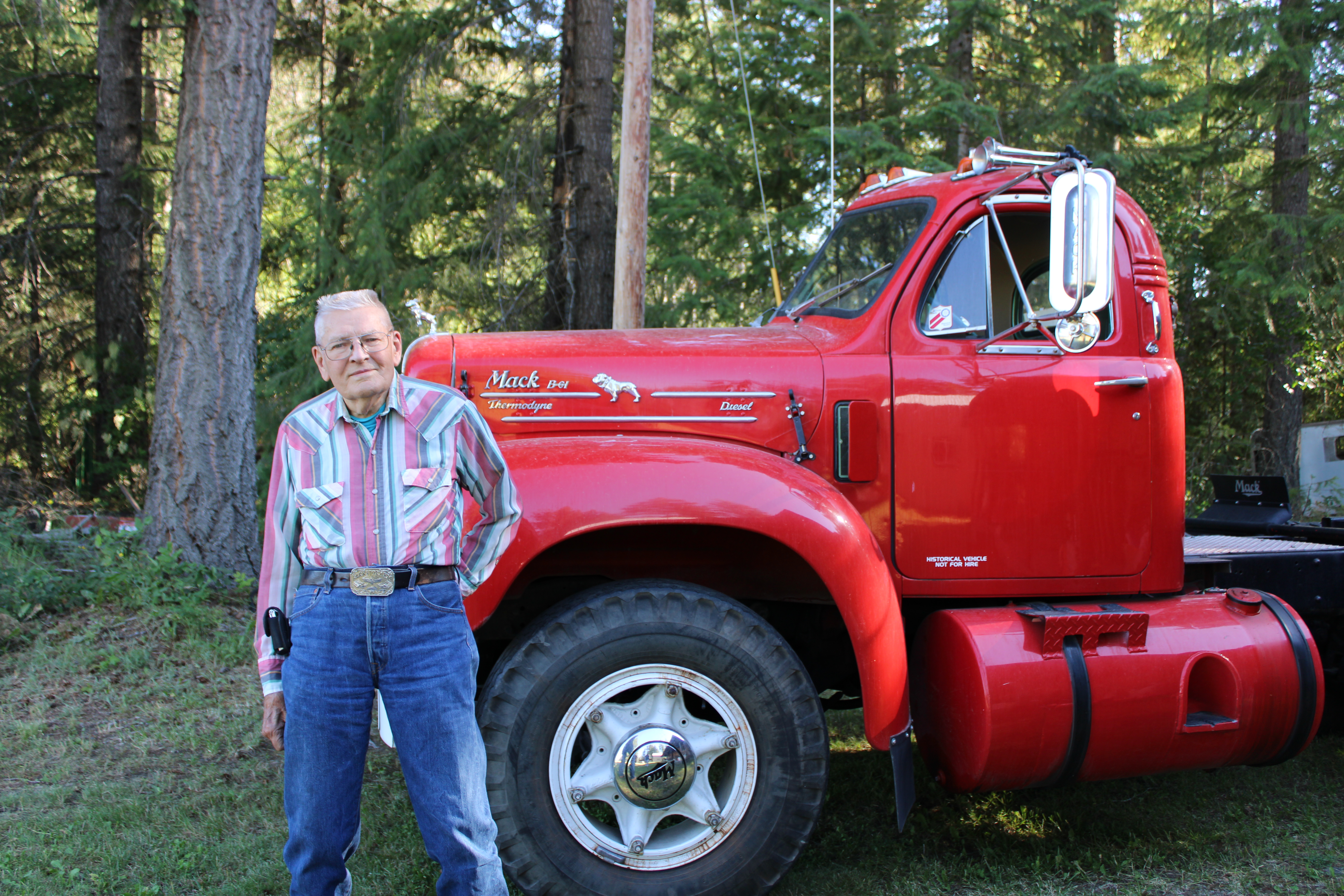 Gene Soper standing beside a classic red Mack B61 Thermodyne Diesel semi-truck, set against a backdrop of green trees.