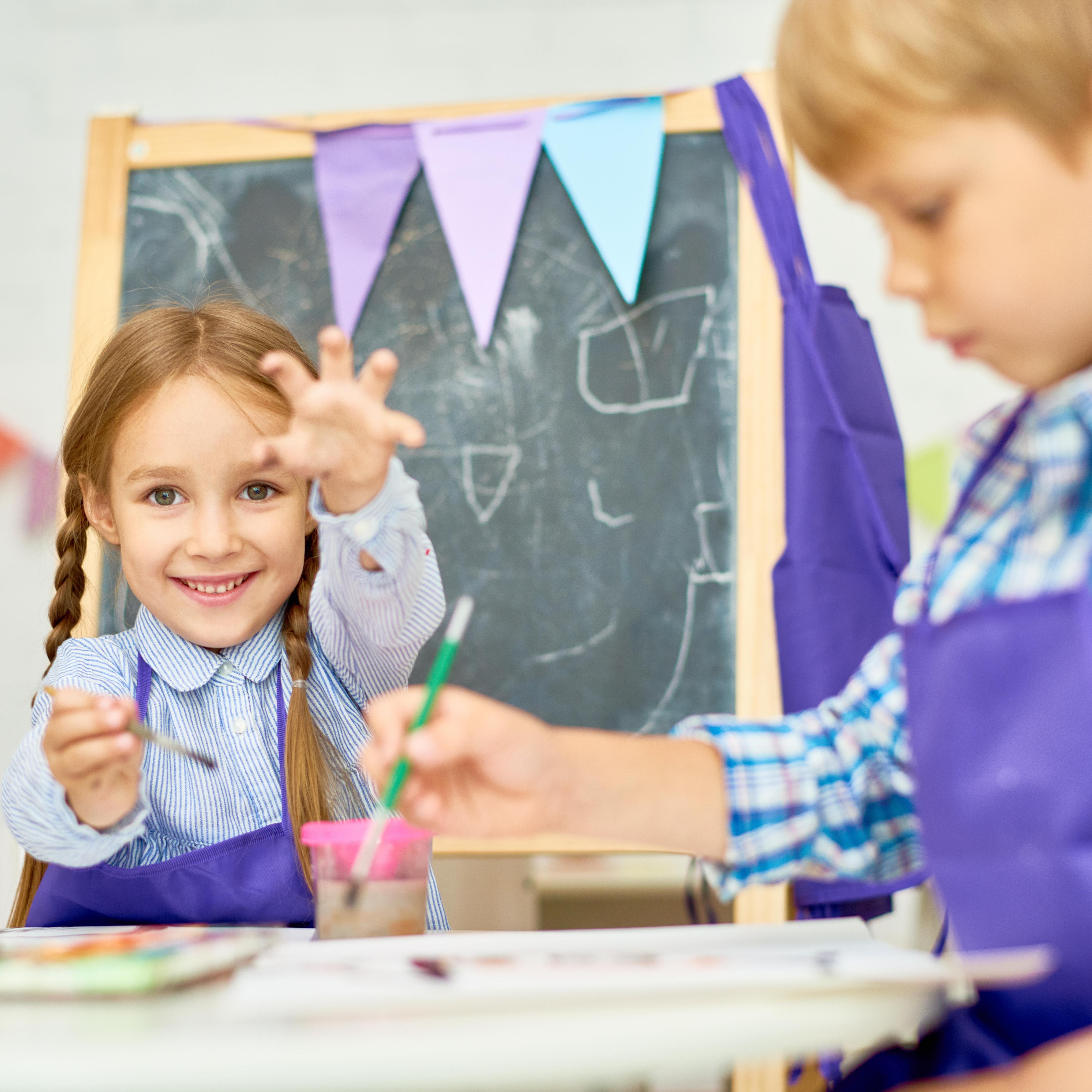 A boy painting with a brush and a girl showing her painted hand behind him, with art supplies on the table and a chalkboard in the background.